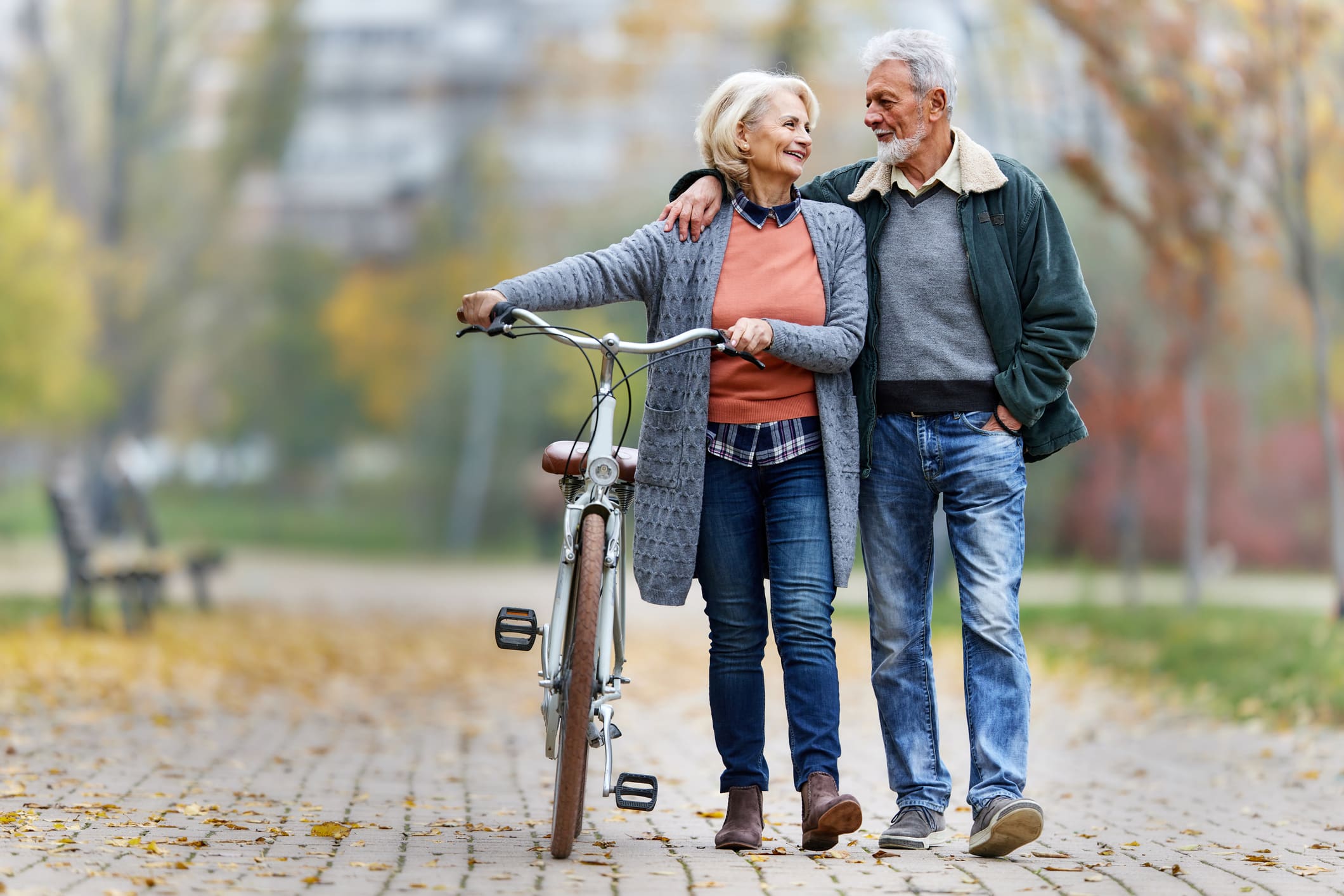Happy mature couple talking while walking embraced in autumn day. The woman is holding a bike alongside her as they walk.