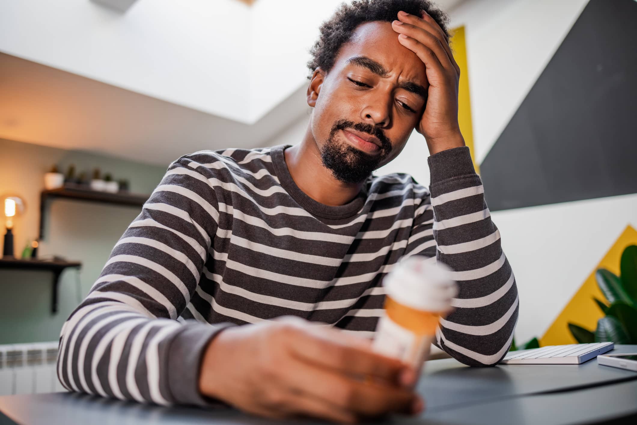 Close-up of a young Black man holding a pill bottle, frowning with his head in his hand.